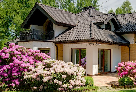 Haus mit Garten, blühenden Sträuchern und Terrasse an einem sonnigen Tag.
